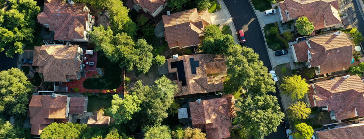 Aerial view of suburban neighborhood with multiple homes featuring rooftop solar panels on terracotta tile roofs surrounded by trees